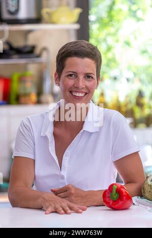 Junge brünette Frau mit kurzen Haaren, die sich auf einem Tisch stützt, Küchenutensilien im Hintergrund. Stockfoto