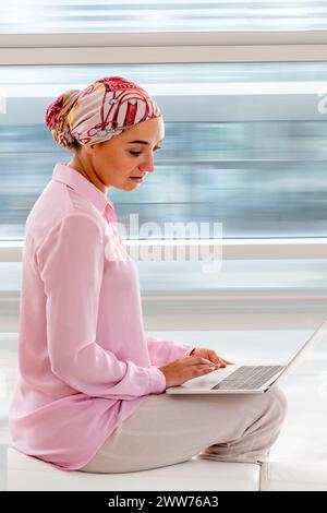 Junge Frau im Profil, die ein Kopftuch trägt, aktiv, mit einem Laptop. Stockfoto