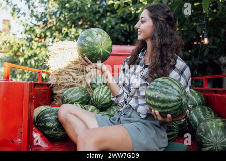 Charmante lächelnde Frau in lässigen Kleidern wirft Wassermelone draußen. Jungbauern mit lockigen Haaren sammeln frische, köstliche Beeren und Früchte. Ernte Stockfoto