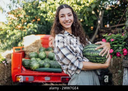 Charmante lächelnde Frau in lässigen Kleidern wirft Wassermelone draußen. Jungbauern mit lockigen Haaren sammeln frische, köstliche Beeren und Früchte. Ernte Stockfoto