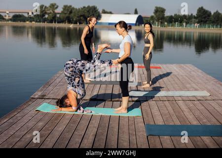 Yoga-Kurs am Morgen auf einem hölzernen Pier in der Stadt. Junge Frauen stehen in einem Kopfständer auf Matten. Der Kursleiter hilft bei der Übung. Eine Gruppe von Frauen A Stockfoto