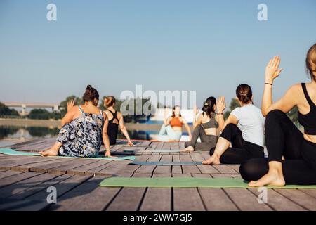 Yoga-Kurs am Morgen mit Lehrer am City Pier. Junge Frauen sitzen in der Pose eines Fischkönig und machen Ardha Matsyendrasana am Flussufer. Gruppe von Mädchen i Stockfoto