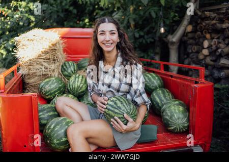 Charmante lächelnde Frau in lässigen Kleidern wirft Wassermelone draußen. Jungbauern mit lockigen Haaren sammeln frische, köstliche Beeren und Früchte. Ernte Stockfoto