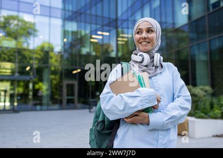 Ein junger Mensch mit Hijab und Rucksack, der Bücher hält, während er vor einem modernen Bildungsgebäude steht. Stockfoto