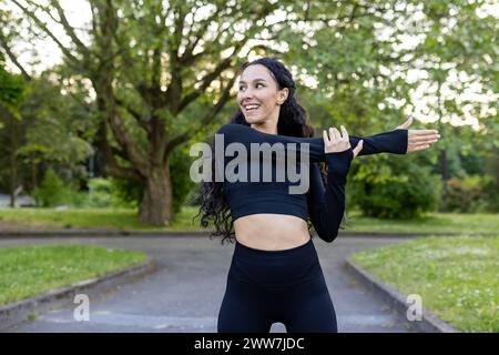 Konzentrierte Frau in Sportbekleidung, die einen Arm dehnt und sich auf ein Workout in einem grünen Park vorbereitet. Stockfoto