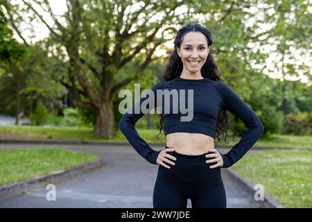 Eine lächelnde junge Frau in Sportbekleidung steht selbstbewusst mit Händen auf Hüften in einer üppigen Parklandschaft und strahlt Fitness und Wohlbefinden aus. Stockfoto