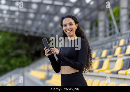 Eine lächelnde junge Frau in Sportkleidung hält ein Smartphone, während sie in einem Sportstadion mit Sitzen im Hintergrund steht. Stockfoto