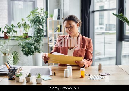 Fokussierte afroamerikanische Ernährungsberaterin, die Umschläge in der Nähe von Nahrungsergänzungsmitteln auf ihrem Schreibtisch überprüft Stockfoto