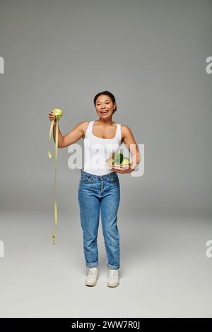 Fröhlicher afroamerikanischer Ernährungsberater mit Apfel und Nahrungsergänzungsmitteln, die Gesundheit vor grauem Hintergrund zeigen Stockfoto