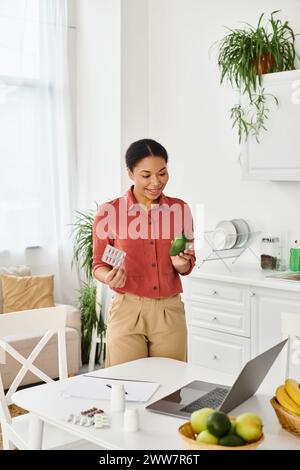 Glücklicher afroamerikanischer Ernährungswissenschaftler hält reife Avocado und gibt Ernährungsratschläge auf dem Laptop in der Küche Stockfoto