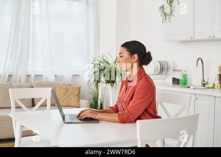 Lächelnde afroamerikanerin, die ihren Laptop benutzt, während sie von zu Hause aus schreibt und arbeitet Stockfoto