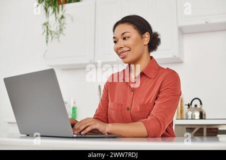 Lächelnde afroamerikanerin, die ihren Laptop benutzt, während sie von zu Hause aus schreibt und arbeitet Stockfoto