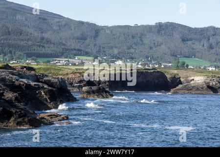 Küstenlandschaft mit felsigen Küsten, turbulenten Gewässern und einer grünen, hügeligen Kulisse mit einem entfernten Dorf Stockfoto