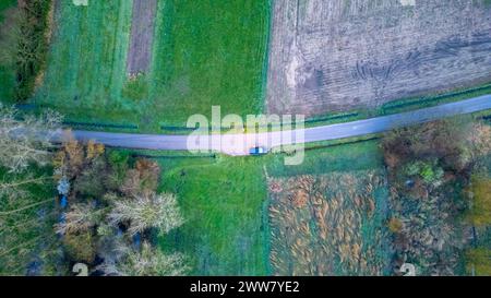 Dieses Luftbild zeigt ein einsames Fahrzeug, das auf einer gewundenen Landstraße fährt, umgeben von einem Flickenteppich aus Feldern und Bäumen, die für die Landschaft charakteristisch sind. Die Ruhe der Szene ist spürbar, während die Natur den Weg und den einsamen Reisenden umhüllt. Reise durch die Landschaft: Ein einsames Auto auf einer Landstraße von oben. Hochwertige Fotos Stockfoto