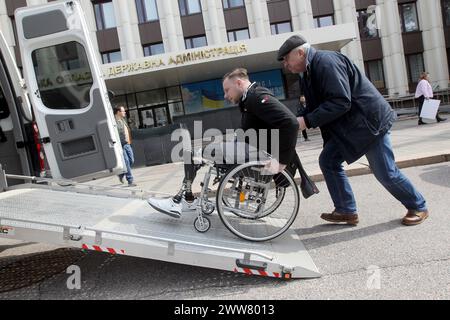 DNIPRO, UKRAINE - 21. MÄRZ 2024 - Ein Veteran in einem Rollstuhl mit prothetischen unteren Gliedmaßen wird nach der Teilnahme an der Präsentation eines neuen Sozial- und Bildungsprojekts der regionalen Militärverwaltung Dnipro Titans.UA, Dnipro, Ostukraine, geholfen, in einem spezialisierten Auto Platz zu nehmen. Stockfoto