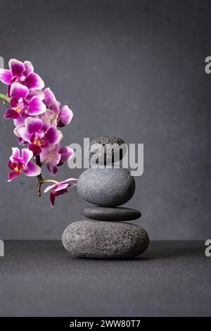 Ein heiterer Stapel glatter grauer Steine, die in einem Zen-ähnlichen Arrangement ausgewogen sind, mit rosa Orchideenblüten, die einen Hauch von Natur verleihen, und mit einer grauen Textur Stockfoto