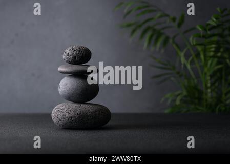 Ein heiterer Stapel glatter grauer Steine, die in einem Zen-ähnlichen Arrangement ausgewogen sind, mit einer subtilen grünen Pflanze, die einen Hauch von Natur verleiht, und einer Textur Grau gegenübersteht Stockfoto