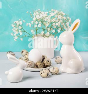 Niedliche Osterkarte mit weißen Keramikhasen, Wachteleiern, zarten weißen gypsophila-Blumen auf weißem Tisch vor türkisfarbenem Hintergrund. Osterdekora Stockfoto