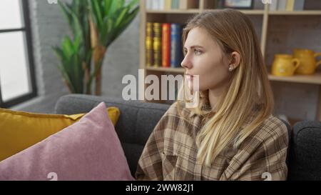 Eine besinnliche junge Frau sitzt drinnen auf einem grauen Sofa, umgeben von bunten Kissen mit einem Bücherregal und einer Zimmerpflanze im Hintergrund. Stockfoto