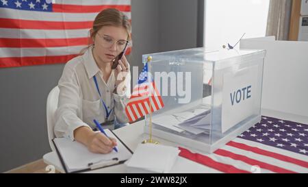 Eine junge kaukasische Frau multitasert, schreibt und spricht am Telefon in einem US-Wahlzentrum mit Fahnen Stockfoto