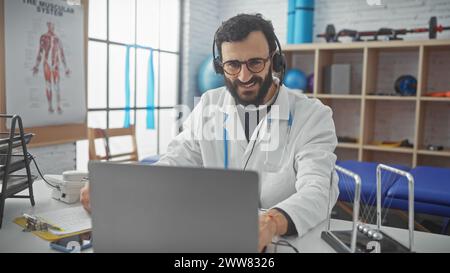 Lächelnder bärtiger Mann mit Labormantel und Kopfhörern, der in der Physiotherapiestelle am Laptop arbeitet Stockfoto