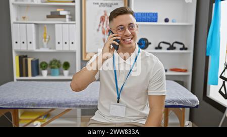 Ein lächelnder junger hispanischer Mann mit Brille und Schlüsselband spricht an einem Telefon in einer modernen Therapieklinik. Stockfoto