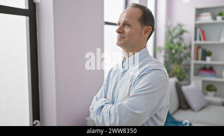 Gutaussehender Mann mittleren Alters, der Glück ausstrahlt, selbstbewusst mit gekreuzten Armen steht und ein fröhliches, lässiges Lächeln in seinem gemütlichen Wohnzimmer zeigt Stockfoto