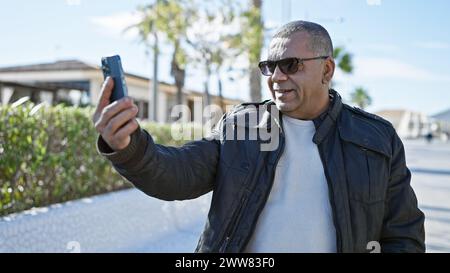 Ein hispanischer Mann mittleren Alters mit Sonnenbrille und Lederjacke macht ein Selfie im Freien in einem sonnigen Stadtpark. Stockfoto