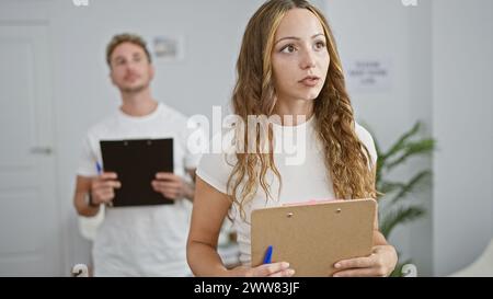 Eine Frau und ein Mann mit Klemmbrettern stehen nachdenklich in einem modernen, weißen Büro-Innenraum, was auf einen professionellen Kontext hindeutet. Stockfoto