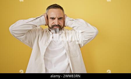 Reifer hispanischer Mann mit runzelndem Bart, der seine Ohren vor gelbem Hintergrund bedeckt. Stockfoto