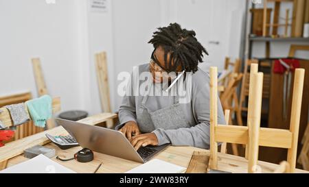 afroamerikanerin mit Dreadlocks, die in einer Werkstatt, umgeben von Holzwerkzeugen und -Projekten, an einem Laptop arbeitet. Stockfoto