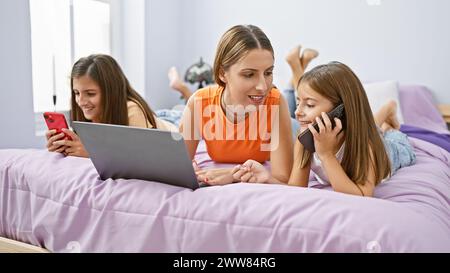 Eine Mutter und ihre beiden Töchter genießen gemeinsam Technologie in einem gemütlichen Schlafzimmer, was Familie, Konnektivität und Zuhause veranschaulicht. Stockfoto