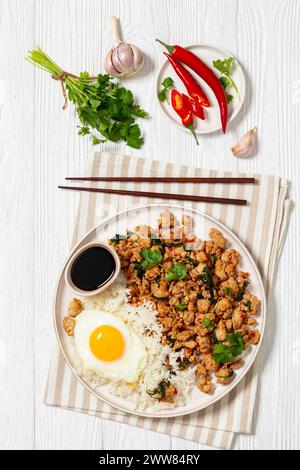 Pad Krapow Gai, Pad Gaprao, Pad kra Pao, Basilikum-Hühnchen-Rührbraten mit Reis und Spiegelei auf Teller mit Essstäbchen auf weißem Holztisch, thai-Küche, Stockfoto