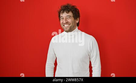 Hübscher junger Mann, der in einem weißen Pullover vor rotem Hintergrund mit stilvollem Bart und zerzausten Haaren lächelt. Stockfoto