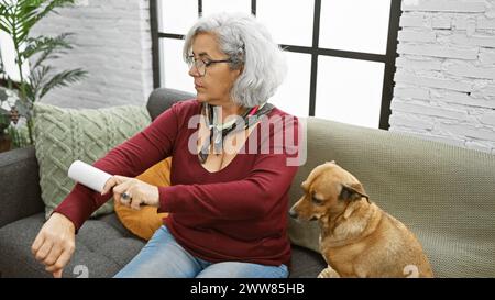 Reife Frau mit grauen Haaren, die einen Fusselroller auf dem Ärmel neben ihrem Hund auf einem Sofa in einem gemütlichen Wohnzimmer benutzt. Stockfoto