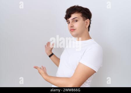 Junger, nicht binärer Mann, der ein lässiges weißes T-Shirt trägt und mit offener Hand zum Eintreten einlädt Stockfoto