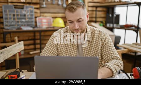 Ein junger kaukasier mit Bart arbeitet an einem Laptop in einer Holzwerkstatt. Stockfoto