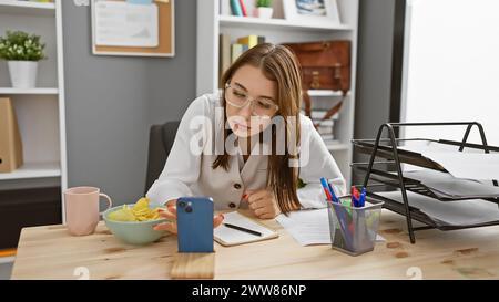 Junge kaukasische russin konzentrierte sich auf die Arbeit in einem modernen Büro, mit Smartphone und Snacks. Stockfoto
