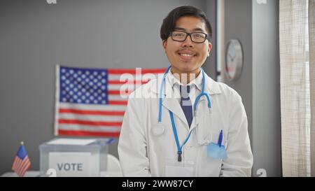 Ein junger asiatischer Mann mit Brille, Laborkittel und Stethoskop, lächelt professionell in einem US-Wahlzimmer mit Wahlurnen und Flagge. Stockfoto