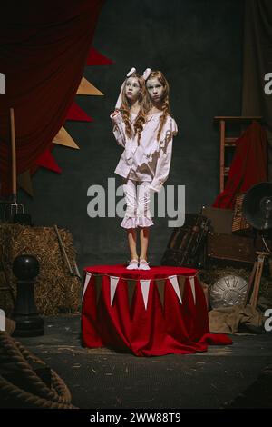 Siamesische Zwillingsmädchen in weißen Vintage-Kostümen mit Make-up-Stand auf der Bühne über dunklem Retro-Zirkus-Hintergrund. Magische Show mit gruseligen Themen Stockfoto