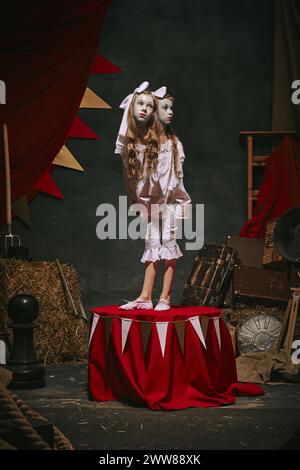 Siamesische Zwillingsmädchen in weißen Vintage-Kostümen mit Make-up stehen auf der Bühne über dunklem Retro-Zirkus-Hintergrund. Stockfoto