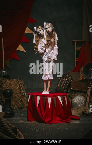 Siamesische Zwillingsmädchen in weißen Vintage-Kostümen mit Make-up stehen auf der Bühne über dunklem Retro-Zirkus-Hintergrund. Stockfoto