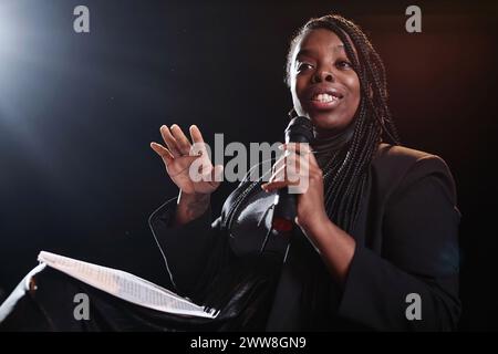 Minimales Porträt einer afroamerikanischen Frau, die auf der Bühne mit einem Mikrofon spricht, mit dunklem Kopierraum im Hintergrund Stockfoto