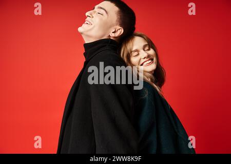 Fröhliches, liebevolles Paar in stilvollen Mänteln, das fröhlich mit geschlossenen Augen vor roter, lebendiger Kulisse lächelt Stockfoto