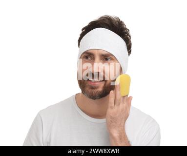 Mann mit Stirnband, der sein Gesicht mit Schwamm auf weißem Hintergrund wäscht Stockfoto
