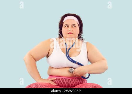Eine junge Frau mit Fettleibigkeit und Verdauungsproblemen hält ein Stethoskop auf ihrem fetten Magen Stockfoto
