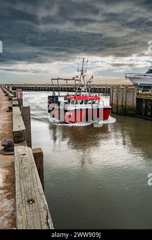 Das Fischerboot kehrt vor einer starken stürmischen Wolkenlandschaft zum Hafen von West Bay zurück. Am späten Nachmittag im Frühling. Kontrastfarben Rot und Grau. Stockfoto