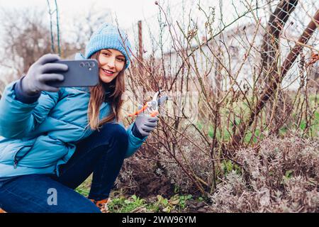 Junger Gärtner Blogger filmt Vlog über das Beschneiden von Rosenstrauch im Frühlingsgarten. Frau, die mit dem Smartphone Inhalte in sozialen Medien erstellt Stockfoto