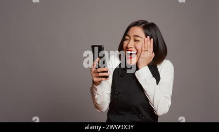 Der asiatische Kellner war zufrieden, nachdem er gute Nachrichten am Telefon gelesen hatte und viele Gäste reserviert hatten. Kellnerin mit Schürze, die mit dem Smartphone Tischreservierungen sieht, arbeitet gerne. Kamera B.. Stockfoto