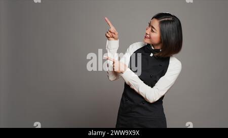 Asiatische Kellnerin zeigt nach oben für eine Werbeanzeige im Studio und gibt die Richtung über ihrem Kopf an, Werbeanzeige zu erstellen. Die Catering-Mitarbeiterin zeigt auf etwas nach oben. Kamera A.. Stockfoto